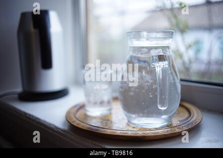 Jug of water and a glass in a tray on the windowsill. Electric kettle in the background. Rural house outside the window Stock Photo