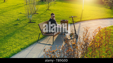 Young attractive man with laptop and modern electric kick scooter sitting at contemporary park at bench. and working Freelancer or business man workin Stock Photo