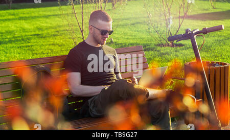 Young attractive man with laptop and modern electric kick scooter sitting at contemporary park at bench. and working Freelancer or business man workin Stock Photo