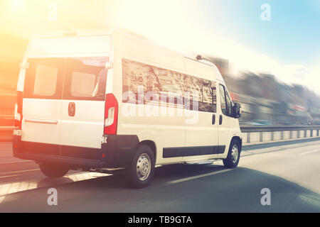 Unrecognizable small passenger van hurry up on highway at city street traffic with urban cityscape and sunset sky on background. Charter or shuttle bu Stock Photo