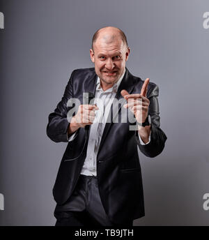 Fun emotional moving happy performer man presenting the show holding microphone in hand and showing the finger the choosing sign on grey color backgro Stock Photo