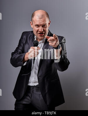 Fun emotional moving happy performer man presenting the show holding microphone in hand and showing the finger the choosing sign on grey color backgro Stock Photo