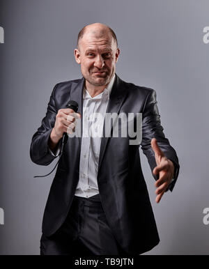 Fun emotional moving happy performer man presenting the show holding microphone in hand and showing the hand the welcome sign on grey color background Stock Photo