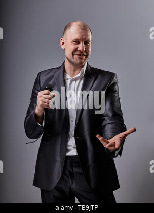 Fun emotional moving happy performer man presenting the show holding microphone in hand and showing the hand the welcome sign on grey color background Stock Photo