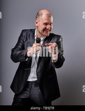 Fun emotional moving happy performer man presenting the show holding microphone in hand and showing the hand the welcome sign on grey color background Stock Photo