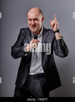 Fun emotional moving happy performer man presenting the show holding microphone in hand and showing the finger with idea sign on grey color background Stock Photo