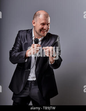 Fun emotional moving happy performer man presenting the show holding microphone in hand and showing the finger the choosing sign on grey color backgro Stock Photo