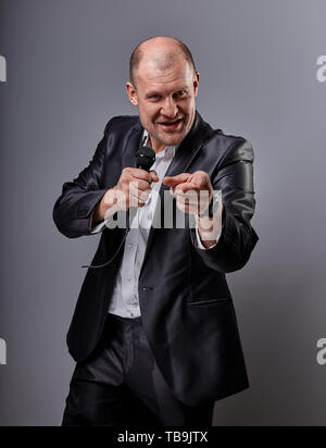 Fun emotional moving happy performer man presenting the show holding microphone in hand and showing the finger the choosing sign on grey color backgro Stock Photo
