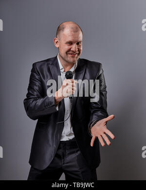 Fun emotional moving happy performer man presenting the show holding microphone in hand and showing the finger the choosing sign on grey color backgro Stock Photo