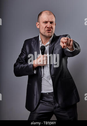 Fun emotional moving happy performer man presenting the show holding microphone in hand and showing the finger the choosing sign on grey color backgro Stock Photo