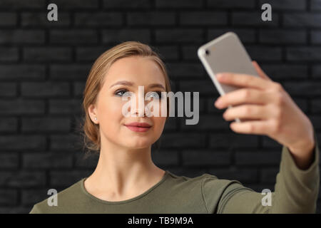 Young woman taking selfie with mobile phone on dark brick background Stock Photo