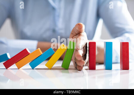 Close-up Of A Businesswoman Hand Stopping Colorful Dominoes From Falling On Office Desk Stock Photo