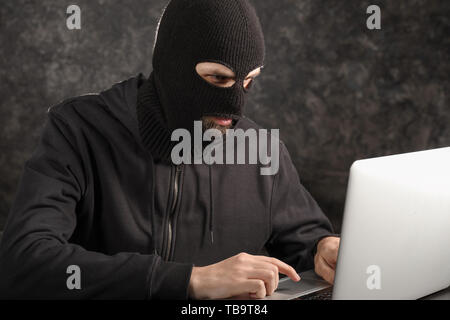 Male hacker in mask using laptop on dark background Stock Photo