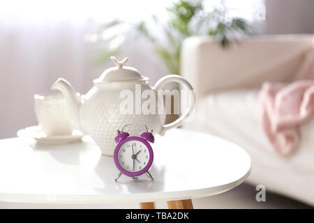 Alarm clock, teapot and cup on table in light room Stock Photo