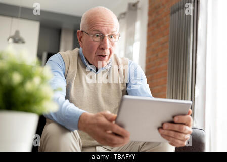 Senior man surprised using a digital tablet while sitting on couch at home Stock Photo