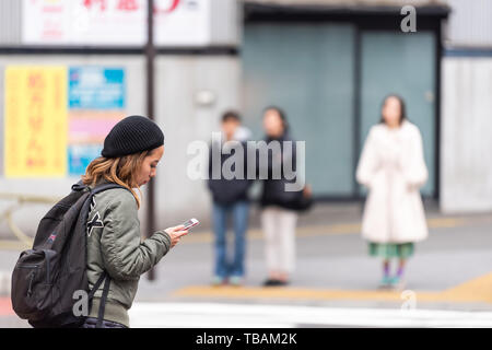 Tokyo, Japan - March 30, 2019: Shinjuku street sidewalk with woman girl waiting to cross road pavement by station building during day with phone Stock Photo