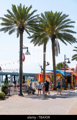 Destin, USA - April 24, 2018: People walking by marina with stores, rental shops and palm trees at Harborwalk village city town on sunny day in Florid Stock Photo