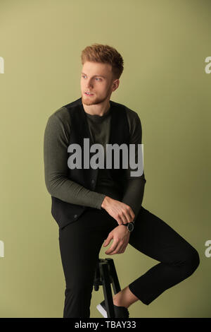 Fashionable young man sitting on stool against color background Stock Photo