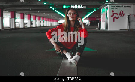 Pretty young woman in red hoodie sitting on car parking. Wearing stylish urban outfit