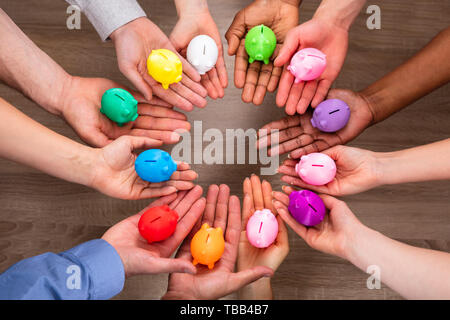 Elevated View Of People's Hand Holding Colorful Piggybanks Over Wooden Desk Stock Photo
