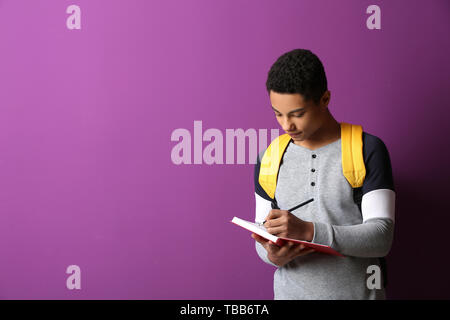 African-American schoolboy writing in notebook on color background Stock Photo