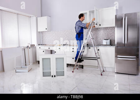 Rear View Of A Male Carpenter Assembling White Cabinet On Wall In Kitchen Stock Photo