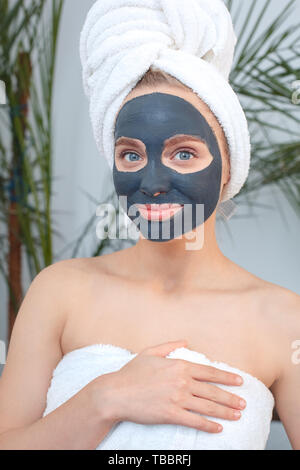 Young woman wearing towel rest at home beauty care standing with a black clay mask on face looking camera smiling happy Stock Photo