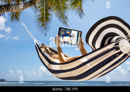 Woman Relaxing On Hammock Over The Sea Looking At Home Security Camera On Digital Tablet Stock Photo