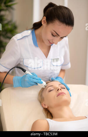 Face lifting. Dark-haired dermatologist using modern equipment while doing face lifting for woman Stock Photo