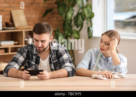 Lazy husband and sad wife sitting at table in kitchen Stock Photo