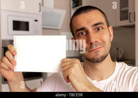 Young man holding and show blank paper notepad with space for text. Kitchen background blurred. Stock Photo