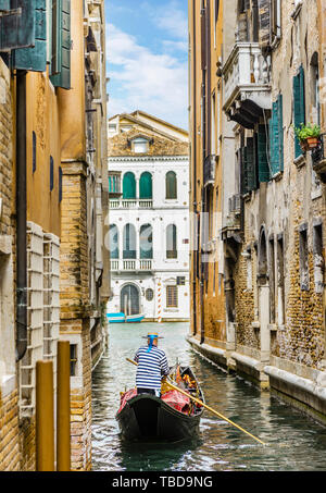 Gondolier in a stripped vest floating on a gondola through a narrow channel among the houses in Venice Stock Photo