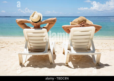 Rear View Of A Young Couple Wearing Hat Relaxing On Deck Chair On Beach Stock Photo
