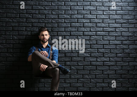 Fashionable young man sitting near dark brick wall Stock Photo