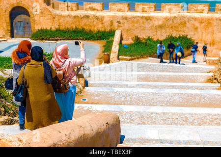 Three Arab girls spend time together in the fresh air, walking in the afternoon along the embankment of the city and taking pictures of the ocean on a Stock Photo