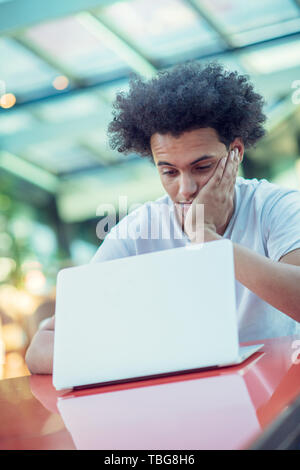 Serious Afro American male student in T-shirt sitting at cafeteria drinking takeaway coffee working at his project using laptop Stock Photo