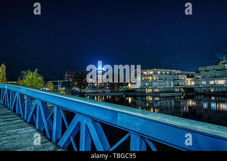 nightshot of the wooden blue bridge crossing the water of Humboldt harbour in Berlin Tegel with illuminated buildings in the background Stock Photo