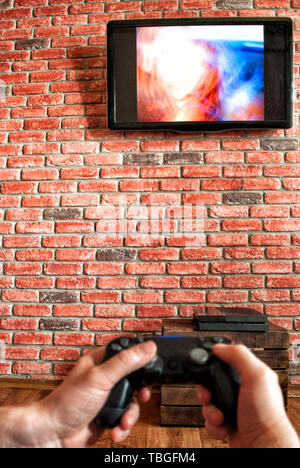 brick wall with a TV and remote control from the game console in the hands in the foreground. Focus on the wall with TV Stock Photo