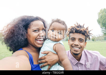 Happy african family taking a selfie with mobile phone in a public park outdoor - Mother and father having fun with their daughter Stock Photo