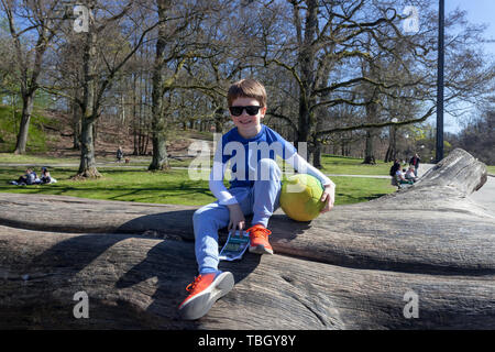 6 year old boy sitting on a tree trunk, holding a ball and a free official map of the park in Slottsskogen Park Gothenborg, Sweden. Stock Photo