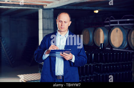 Confident male winemaker working in wine cellar, taking notes on clipboard Stock Photo