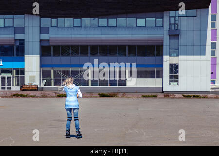 A young blonde girl in a blue jacket is roller-skating around the square for walks and rest on a clear spring day. Stock Photo