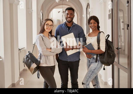 Front view of three best friends Africans and Caucasian standing on corridor of university with backpacks and notes in hands. Students going together on lecture in university. Stock Photo