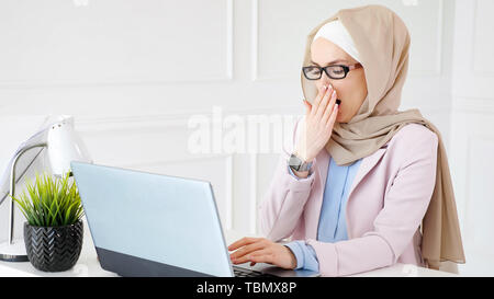 Weary muslim woman in glasses and hijab at the end of working day is typing on laptop and yawning at her workplace. Stock Photo