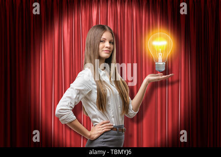 Young brunette business woman holding neon yellow light bulb on red stage curtains background Stock Photo