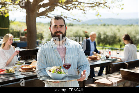 Hospitable young owner of cosy country restaurant in open air meeting guests with serving tray in hands Stock Photo