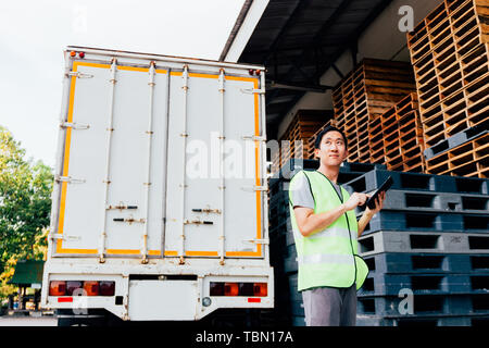 Young Asian male logistic warehouse distribution business entrepreneur using tablet. He surrounded by plenty of pallets and truck in shipping cargo. Stock Photo