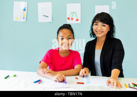 Cheerful Asian woman smiling and looking at camera while teaching African American girl to draw with crayons at table in art school Stock Photo