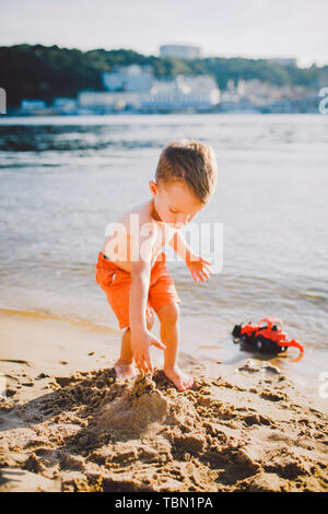 Subject construction and heavy industry. Abstraction child boy playing on the sand near the river in the summer toy red tractor model, excavator Stock Photo