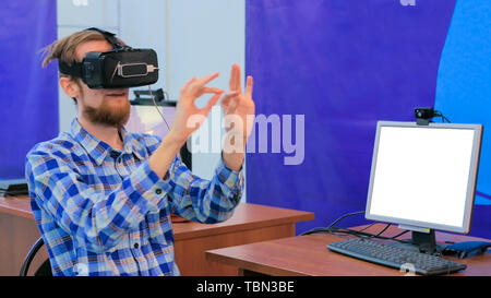 Young man using virtual reality headset in front of white blank monitor Stock Photo